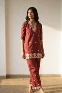 Woman wearing a red traditional outfit with white patterns against a plain wall.