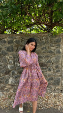 Woman in a purple patterned dress standing against a stone wall with greenery.