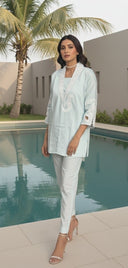 Woman in light blue top and white pants standing by a pool with palm trees in the background