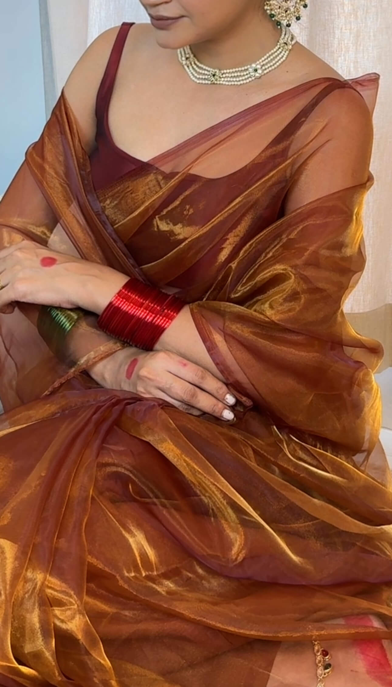 Woman wearing orange, brown organza saree with gold shine, glowing under sunlight. Paired up with complimentary minimalistic bangles and necklace.