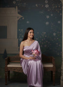 Woman in a lavender saree holding a pink flower, sitting on a wooden bench against a decorative wall, a minimal saree look.