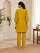 Woman wearing a mustard yellow outfit in a room with plants and a wooden table.