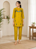Woman wearing a yellow embroidered outfit standing in a room with plants and a wooden table.