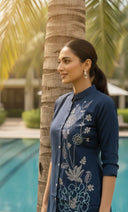 Woman in a blue embroidered outfit standing by a pool with palm trees in the background