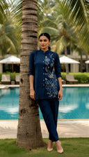 Woman in a blue embroidered outfit standing by a palm tree by a poolside.