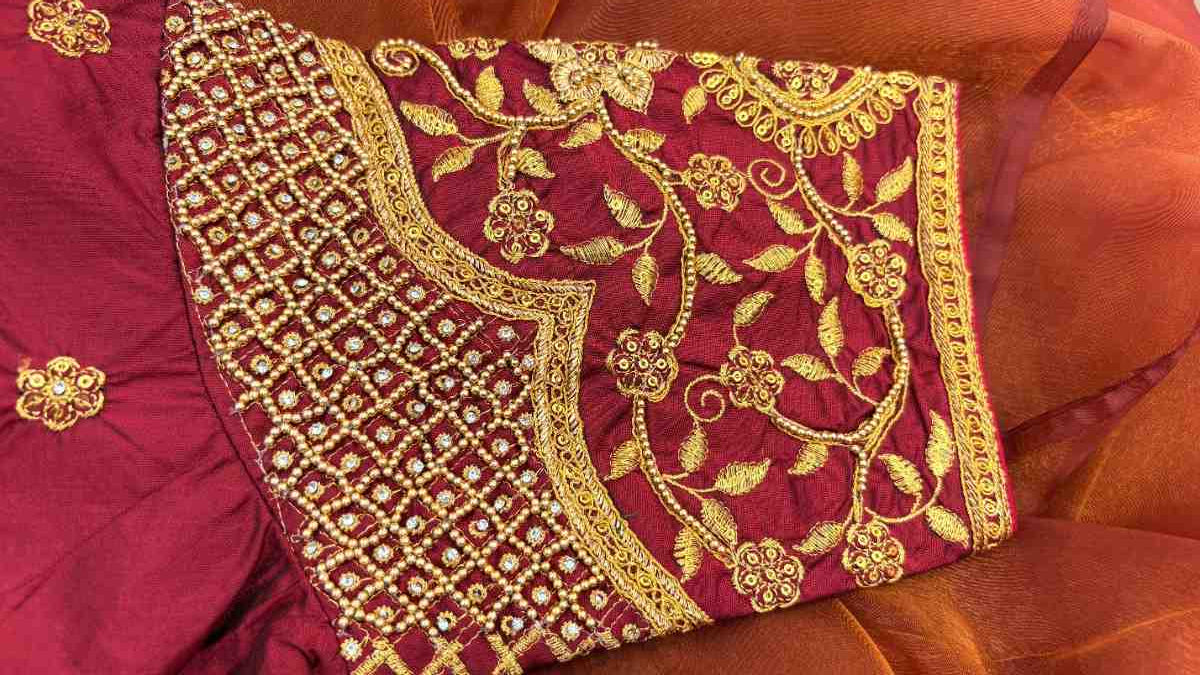 Close-up of the sleeve of Label Anaira’s Noor-e-Zari Blouse showcasing intricate gold Aari embroidery on a rich maroon silk base. The design features hand-embroidered floral and paisley motifs highlighted with zari and resham threads, bordered by an ornate cuff that glimmers with traditional craftsmanship. The fine detailing and dense stitchwork reflect artisanal skill, making it a luxurious choice for brides and festive occasions.