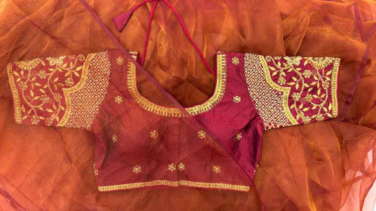Styled look of the Noor-e-Zari Blouse paired with a wedding or festive saree. Deep maroon silk blend with heavy gold embroidery, floral and paisley motifs, and thick neckline borders create a luxurious, regal, and statement-making ensemble