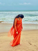 Woman in an orange saree standing on a beach with ocean waves in the background