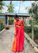 Woman in Pink Pink Pink Tissue Saree standing on a stone path with greenery and a colorful structure in the background.