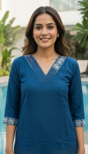 Woman wearing a blue dress with decorative trim standing by a pool.