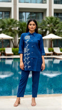 Woman in a blue embroidered outfit standing by a pool with a building and palm trees in the background.