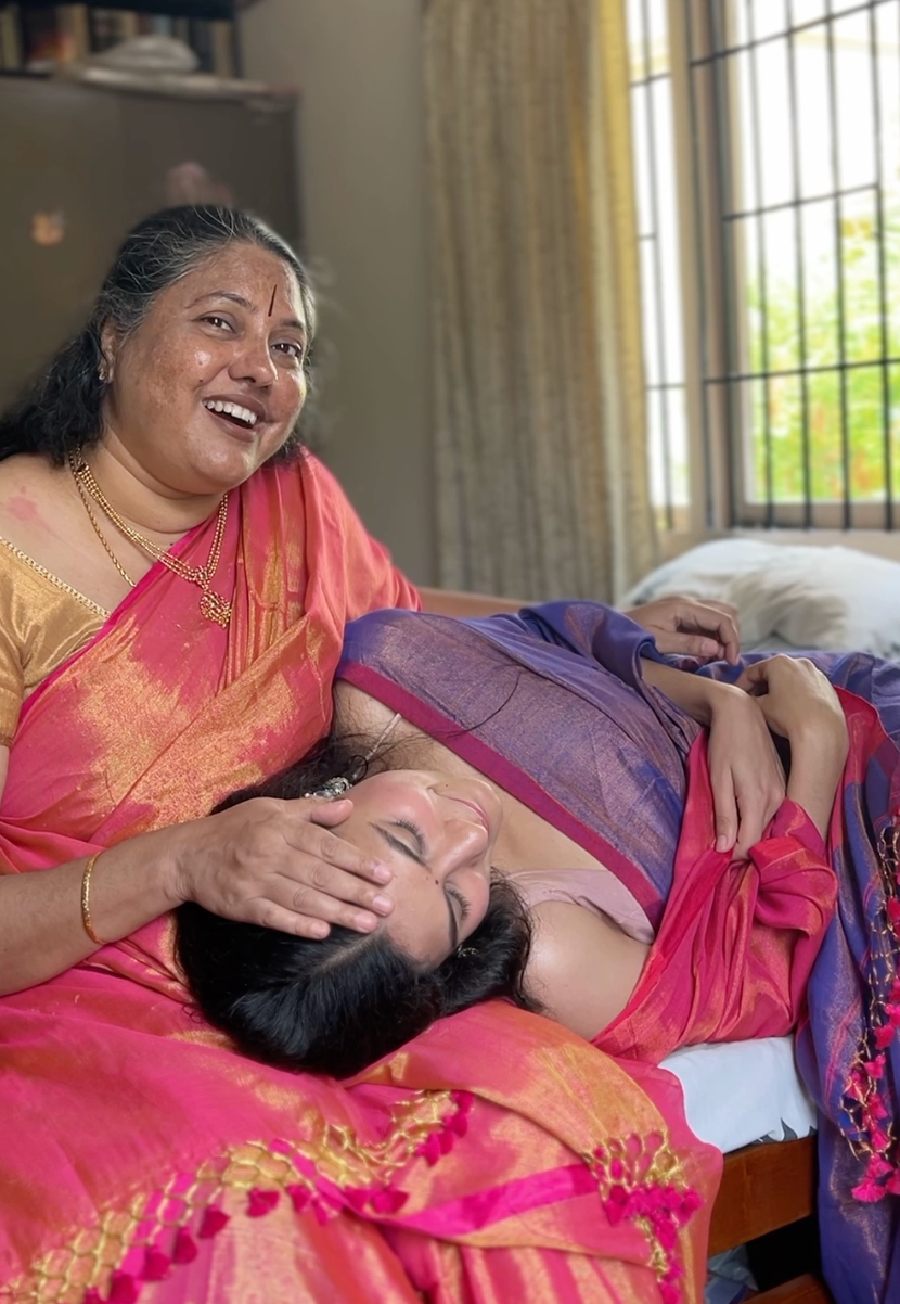 Two women in traditional sarees, one sitting and one lying down, wearing linen tissue sarees, pink and purple shades with shine under light.