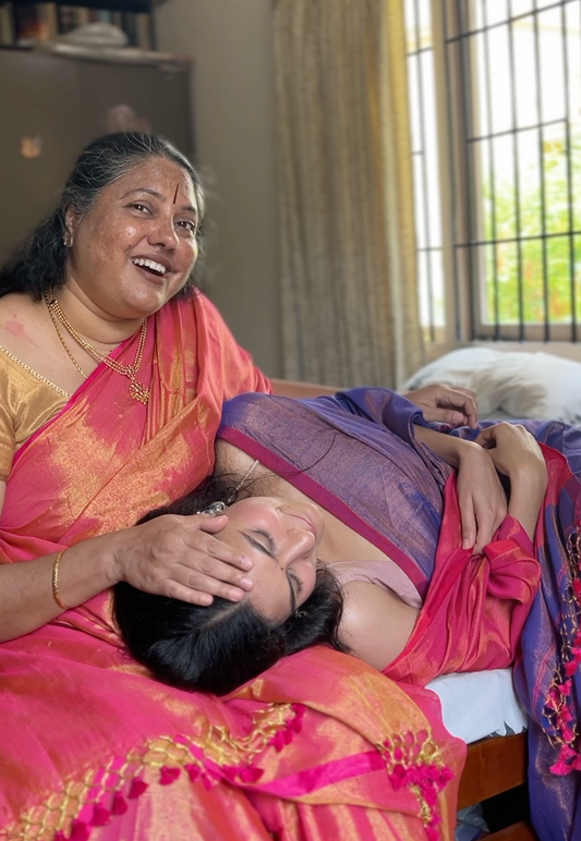 Two women in traditional sarees, one sitting and one lying down, wearing linen tissue sarees, pink and purple shades with shine under light.