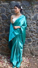Woman in a teal satin saree standing against a stone wall.