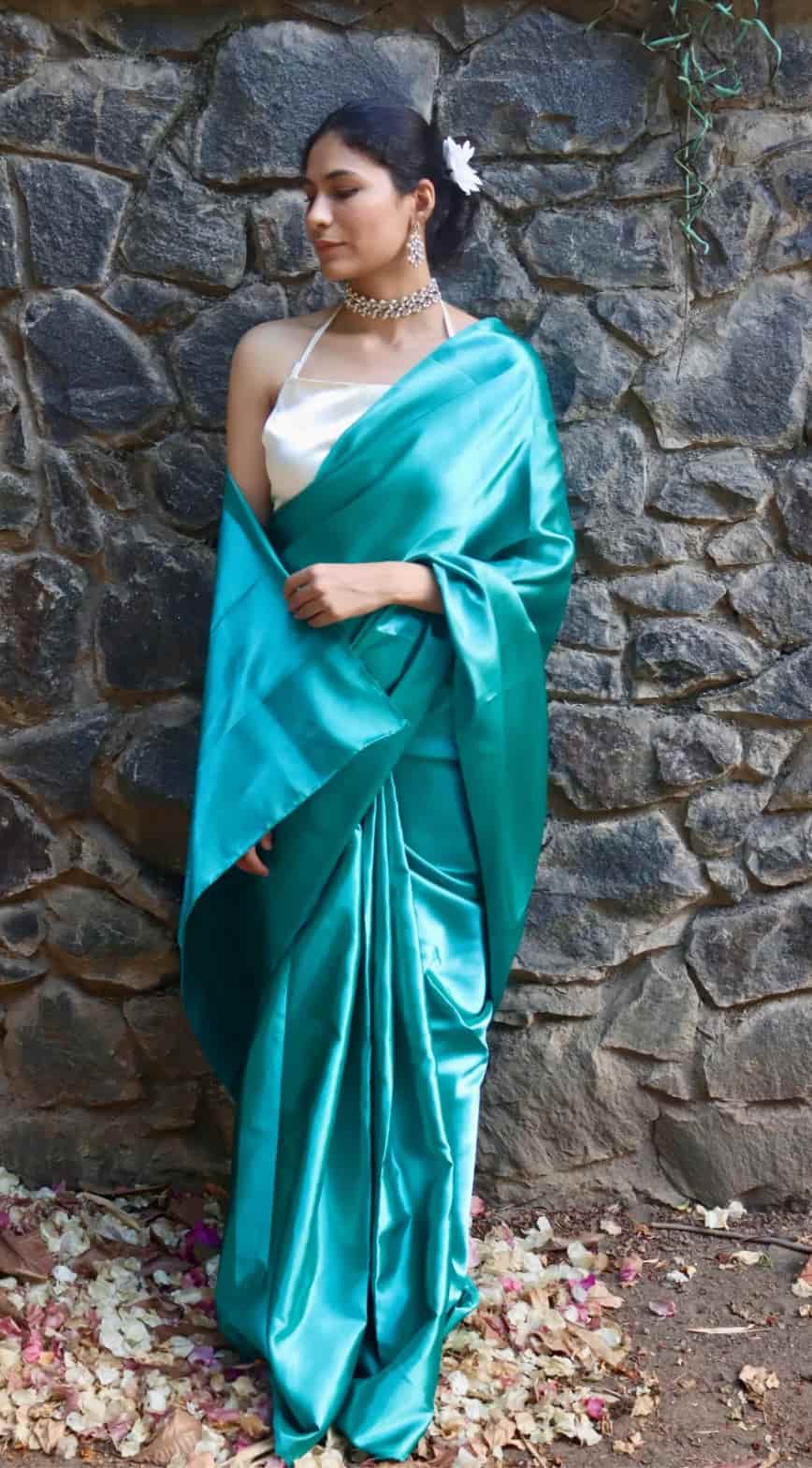 Woman in a teal satin saree standing against a stone wall.