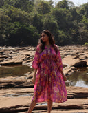 Woman in a pink purple free size dress standing by a river with trees in the background