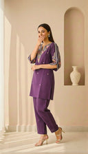 Woman in a purple outfit standing in a well-lit room with a vase on a shelf.