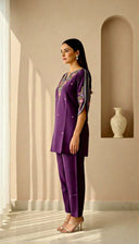 Woman in a purple outfit with embroidery standing in a sunlit room.